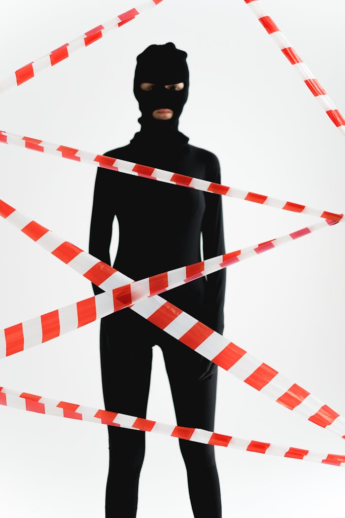 Silhouette of a person in black outfit and balaclava surrounded by red and white warning tape on white background.